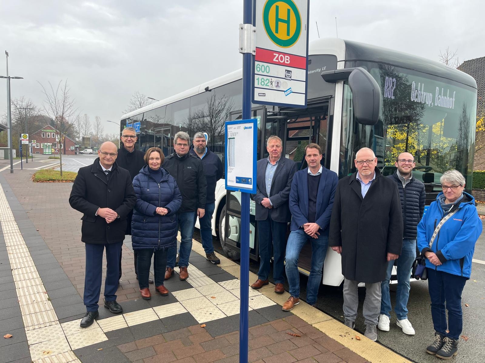 Sie sehen: Eine Personengruppe an einer Bushaltestelle mit Bus im Hintergrund.