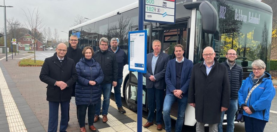 Sie sehen: Eine Personengruppe an einer Bushaltestelle mit Bus im Hintergrund.
