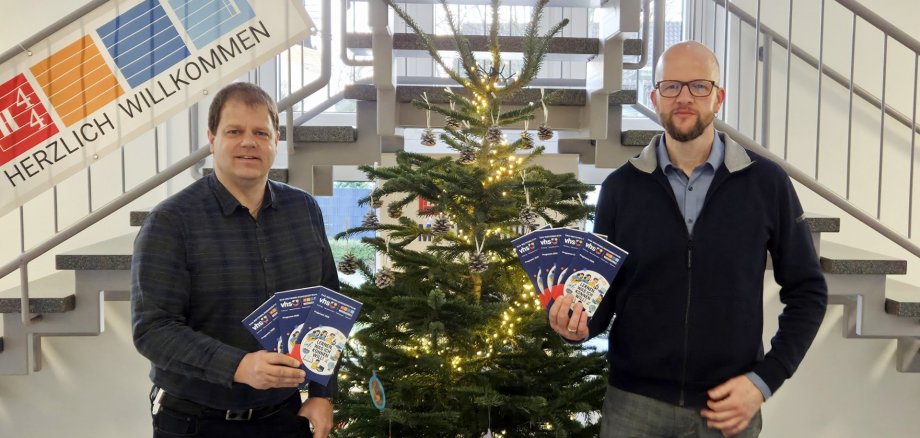 Sie sehen: Zwei Personen neben einem Weihnachtsbaum (Mitte), die Programmhefte präsentieren
