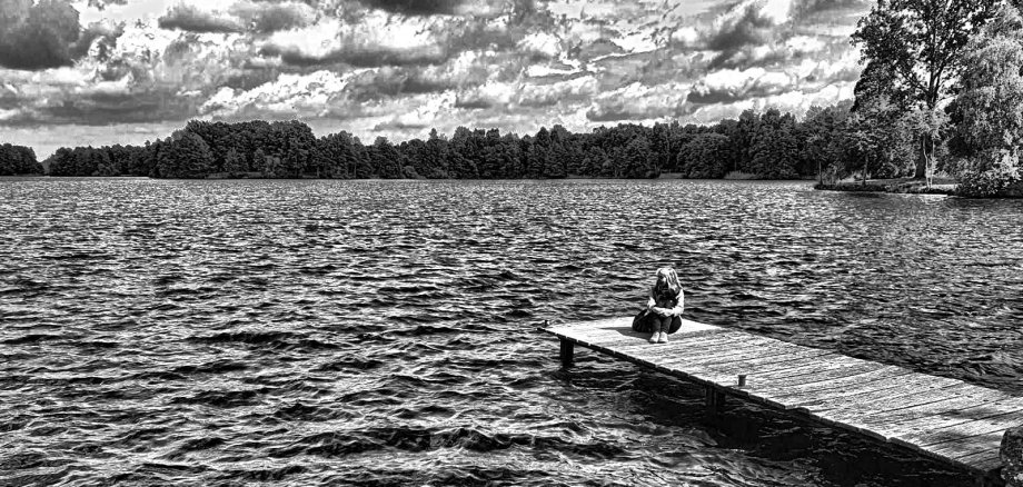 Sie sehen: eine Seenlandschaft und eine sitzende Frau auf einem einsamen Steg (Schwarz-Weiß-Foto)