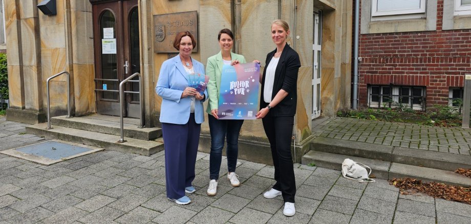 Sie sehen: Drei Personen vor dem Ochtruper Rathaus mit Plakat des Kulturfestivals (Bürgermeisterin Christa Lenderich, Ina Schulze-Bilk und Uta Wienefoet von der Volkbank Ochtrup-Laer eG).