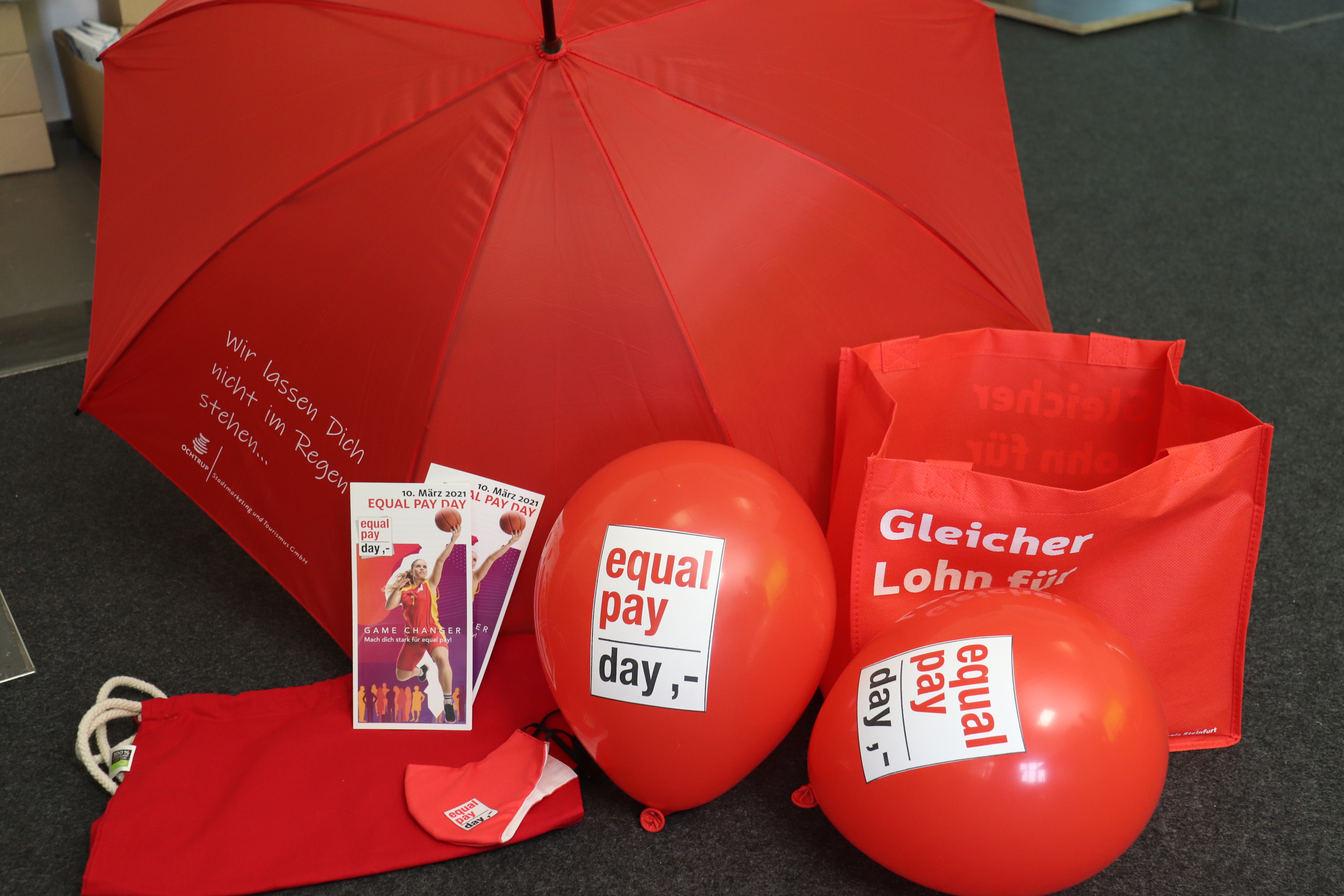Sie sehen: rote Werbematerialien des Equal Pay Day (Taschen, Luftballons, Regenschirm, Flyer)