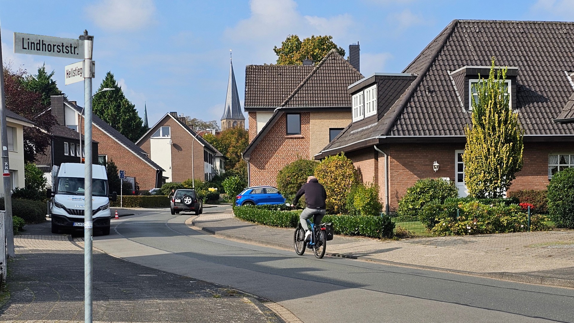 Sie sehen: Kreuzungsbereich von zwei Straßen mit den Straßenschildern, einem Radfahrer und parkenden Autos.