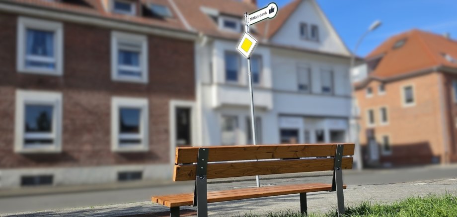 Sie sehen: Eine Sitzbank an einer Straße, davor ein Mast mit zwei Verkehrsschildern; eins trägt die Aufschrift "Mitfahrbank"