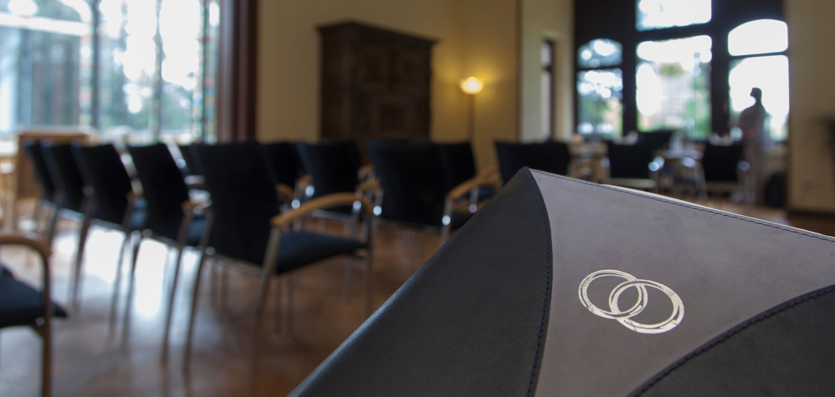 Sie sehen: einen Saal in der Villa Winkel mit Stuhlreihen, im Vordergrund ein Buch mit zwei Trauringen