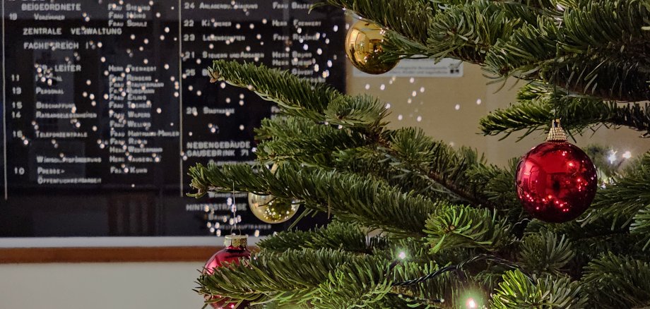 Sie sehen: Weihnachtliche Tannenbaumzweige und Lichter vor einer Anzeigentafel im Rathaus
