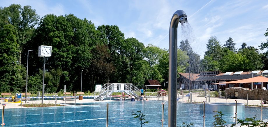 Sie sehen: ein Schwimmbecken im Freibad mit Brause, Wasserbrücke, Uhr und Sanitärgebäuden im Hintergrund